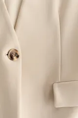 Blazer de punto grueso en color beige, con diseño sin cuello, escote en V, hombreras y cintura entallada. Cuenta con cierre de un botón frontal y bolsillos delanteros con solapa.