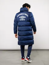 Parka larga acolchada color azul marino con capucha, cierre frontal con botones a presión y bolsillos laterales. Presenta el logo de Umbro y el escudo del Club Nacional de Football bordados en el pecho.