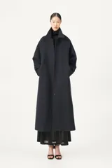 Tapado largo de corte amplio en color azul marino, confeccionado en una mezcla de poliéster y lana. Presenta un diseño con cuello camisero, cierre frontal mediante botones y puños con cintos regulables para un ajuste personalizado.