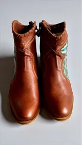 Botas de cuero color marrón tierra con taco medio, detalles de costuras artesanales en el borde superior y un aplique lateral con diseño geométrico pintado en tonos turquesa y cobrizo.