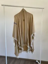 Ruana de tela fina color beige con textura sutil, diseño abierto y bordes decorados con flecos blancos.