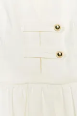 Top de color blanco con escote en pico, mangas cortas abullonadas y bajo con diseño peplum. Presenta detalle de botones dorados en la parte frontal y cierre lateral con cremallera oculta.