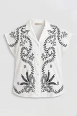 Camisa blanca de manga corta con cuello solapa y cierre frontal de botones, adornada con bordados negros de motivos florales y vegetales.