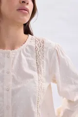 Blusa casual de color crudo con mangas largas abullonadas, cuello redondo y cierre frontal con botones. Presenta delicados detalles de encaje y pliegues en la parte frontal.