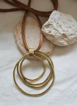 Colgante con dije de tres círculos concéntricos de metal dorado, unidos por una pieza rectangular. Cordón fino de cuero marrón.