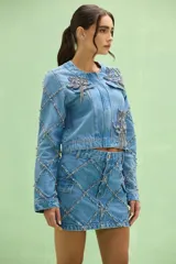 Chaqueta denim de mujer, confeccionada en jean azul claro, con cuello redondo y cierre frontal. Destaca por apliques bordados florales con pedrería y flecos de cristales en la parte frontal, además de detalles de brillos y costuras decorativas a lo largo de las mangas.