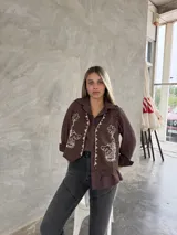 Chaleco marrón de corte corto con bordados florales en color crema en el frente y cierre frontal con botones. Presenta un diseño estructurado con cuello camisero.