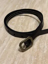Cinturón fino de cuero negro con pespuntes en contraste y hebilla metálica de diseño hexagonal en tono bronce envejecido.