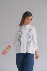 Blusa blanca de corte recto con cuello mao y volado pequeño, cierre frontal con botones y mangas largas con puños elásticos. Presenta un delicado bordado floral en color azul en la parte frontal.