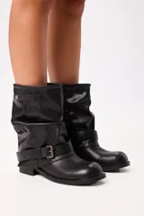 Bota de caña media estilo slouchy en color negro, confeccionada en material sintético con acabado liso. Presenta un diseño holgado en la caña, puntera redondeada, suela plana y una tira decorativa con hebilla metálica ajustable en la zona del tobillo.