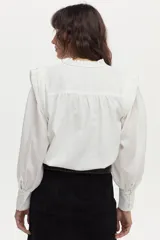 Blusa blanca de tela liviana con bordado detallado en el frente, cuello alto con volado y escote en V con botones. Tiene mangas largas abullonadas con puños amplios y detalles estructurados en los hombros.
