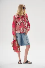 Neceser de lona estampada con diseño floral en tonos rosados y marrones. Presenta bordes en cuero ecológico, cierre metálico superior y un aplique decorativo en forma de caracola dorada. Incluye una correa lateral para llevar en la mano.