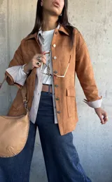 Chaqueta de gamuza color camel, con corte recto, cuello camisero, cierre frontal con botones metálicos y bolsillos frontales con solapa.