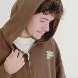 Canguro color marrón con capucha, cierre frontal parcial, bolsillo canguro y cordón ajustable en el dobladillo. Tiene un estampado blanco en el pecho con el texto "PULAU DESIGN DEPT." y un parche amarillo en la manga izquierda.