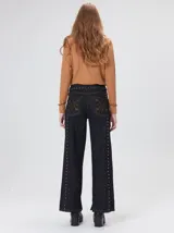 Jeans de corte ancho (wide leg) y tiro medio, confeccionados en denim de algodón negro con costuras contrastantes en color ocre. Presenta múltiples botones metálicos frontales y tachas metálicas aplicadas a lo largo de los laterales y la pretina.