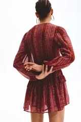 Vestido corto de manga larga confeccionado en tejido transparente, con estampado estilo paisley en tonos borravino y rosado. Presenta cuello redondo con abertura frontal y cordones para atar, cintura elástica y falda con volados en capas.
