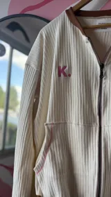 Campera de algodón premium color crudo con detalles en marrón y rosa. Presenta un cierre frontal, cuello redondo, mangas largas acampanadas y un logo bordado en el pecho.