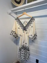 Vestido corto color crudo con bordado floral negro, escote en V, mangas 3/4 acampanadas y cintura ajustable con cordón.