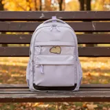 Mochila escolar de color lila con diseño funcional, cuenta con un compartimento principal amplio, un bolsillo frontal con ventana transparente decorada con un parche de capibara y un bolsillo adicional en la parte inferior. Incluye cierres con tiradores de cuerda y detalles en color marrón en la base.