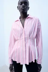 Camisa de popelina con diseño a rayas verticales rosadas y blancas, cuello solapa con escote en V, manga larga y detalle de pliegues marcados en la cintura. Cierre frontal con botones ocultos.