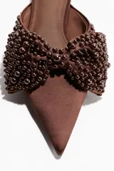 Mules de satén color marrón, con punta fina y una moña grande decorativa en el empeine adornada con cuentas perladas del mismo tono.