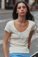 Camiseta efecto doble en color crudo y blanco, de manga corta y corte slim fit. Cuenta con escote redondo, cierre frontal parcial con botones a presión y diseño de capas superpuestas.