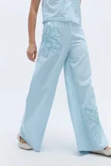 Pantalón de corte amplio y fluido, color celeste, confeccionado en lino. Presenta un diseño con bordados artesanales de flores en relieve en la parte superior del muslo y en la parte inferior de la pierna.