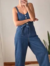 Mono largo de denim azul con tirantes anchos, escote redondo y cierre frontal de botones claros. Cuenta con costuras en hilo tostado y un lazo ajustable en la cintura.