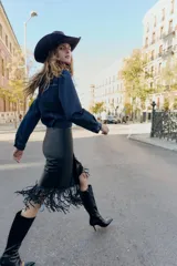 Falda midi negra con efecto piel, diseño cruzado en el delantero y flecos en el bajo. Cierre lateral con cremallera oculta.