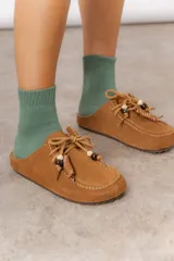 Zueco de cuero gamuzado color tostado, con cordones decorativos adornados con cuentas de madera y piezas envueltas en cuero. Diseño tipo mocasín con costuras visibles en la parte superior y abierto en la parte trasera.