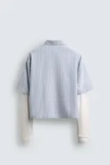 Camisa de corte relajado con diseño de rayas verticales en tonos azul y blanco. Presenta cuello solapa, cierre frontal de botones y un detalle de texto bordado en color rojo en el pecho. Cuenta con un diseño de manga larga con efecto doble, combinando la manga corta de la camisa con una manga larga de tejido tipo waffle en color crudo.