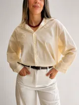 Camisa de corte oversize color beige claro, con mangas largas y cuello camisero clásico. Presenta delicados bordados florales en relieve en la parte superior del frente y abotonadura frontal con botones blancos.