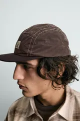 Gorra de visera plana, color marrón oscuro, confeccionada en tejido técnico con costuras contrastantes en blanco. Presenta un parche frontal con un diseño a cuadros y texto.