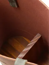 Matera de cuero con diseño bicolor en tonos marrón y verde oliva, con correas largas para hombro y detalles de remaches metálicos. Incluye logo de la marca grabado en el frente.