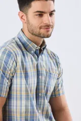 Camisa sport de manga corta con estampa a cuadros en tonos celestes, amarillos y blancos, con cuello camisero abotonado y bolsillo frontal en el pecho.