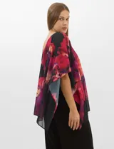 Enterito de seda fría con pantalón corte palazzo de pierna ancha. Presenta un diseño de un hombro descubierto con tirante fino y una sobrecapa asimétrica de gasa estampada con motivos florales en tonos rojos y fucsia sobre fondo negro que cae desde el hombro derecho.
