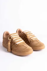 Zapatillas urbanas color tostado con textura geométrica en el cuerpo y detalles de gamuza en la puntera y laterales. Presentan un diseño distintivo con cordones anchos tejidos en tonos tostado y blanco, y un accesorio metálico con borla de gamuza en el empeine.
