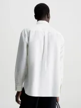 Camisa blanca de manga larga con cuello clásico, cierre frontal con botones, bolsillo de parche en el pecho y logo CK bordado en el bolsillo.