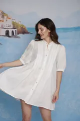 Vestido corto de lino color blanco, con cuello mao, cierre frontal de botones y tablillas decorativas en el pecho. Presenta mangas abullonadas a la altura del codo con terminación elástica y silueta evasé.