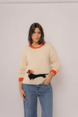 Buzo tejido de lana merino color marfil, con detalles en color naranja en el cuello y los puños. Presenta un diseño estampado de un perro salchicha negro con detalles naranjas en la parte inferior frontal.