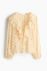 Blusa de gasa arrugada de corte holgado, en tono amarillo claro con estampado floral. Cuenta con escote en V profundo, un amplio volante ribeteado con encaje alrededor del cuello y cordones finos para atar al frente. Mangas largas con puños elásticos.