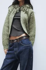 Chaqueta de punto de textura jaspeada en color verde, con cuello redondo adornado con flecos del mismo tejido, manga larga y cierre frontal con corchete metálico.