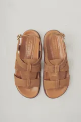 Sandalias planas de gamuza color marrón, con diseño de tiras cruzadas en el empeine y cierre con hebilla metálica dorada en el tobillo. Cuenta con plantilla de cuero y suela de goma con textura.
