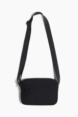 Bandolera negra de nylon con correa ajustable con hebilla deslizante de metal, cierre superior, un compartimento exterior con cierre y dos compartimentos interiores abiertos.