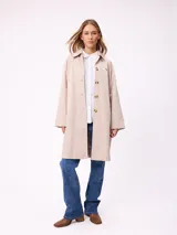Gabardina de corte recto y largo medio, color beige, con cuello camisero, cierre frontal de botones y mangas largas. Presenta un pequeño bordado distintivo en el pecho.