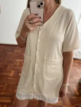 Camisola o bata corta de tejido acanalado color beige claro, con cuello en V, cierre frontal de botones blancos y dos bolsillos delanteros grandes. El ruedo está decorado con un borde de encaje blanco.