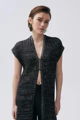 Kimono tejido de punto en color crudo con sutil brillo de lurex. Presenta un diseño sin mangas, corte largo y cierre frontal mediante dos lazos finos. El tejido tiene una textura acanalada vertical.