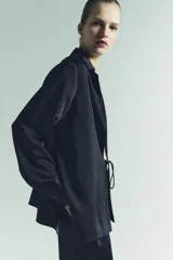 Camisa de manga larga color azul marino, confeccionada en tejido satinado. Presenta cuello solapa, puños anchos y cierre frontal mediante lazada ajustable en el centro.