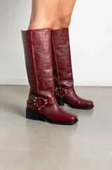 Botas de caña alta en cuero con textura de cocodrilo, color bordó. Presentan un diseño de punta cuadrada, taco bajo y una correa decorativa con argolla metálica y remaches en la zona del tobillo.