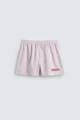 Bóxer tipo short de popelín con estampado de rayas verticales finas en blanco y rosado/rojo. Posee cintura elástica y una pequeña inscripción bordada en rojo en la parte inferior de una de las piernas.